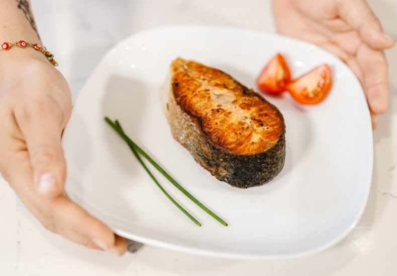 Close-up of grilled salmon served with tomatoes and chives on a plate, held by tattooed hands.